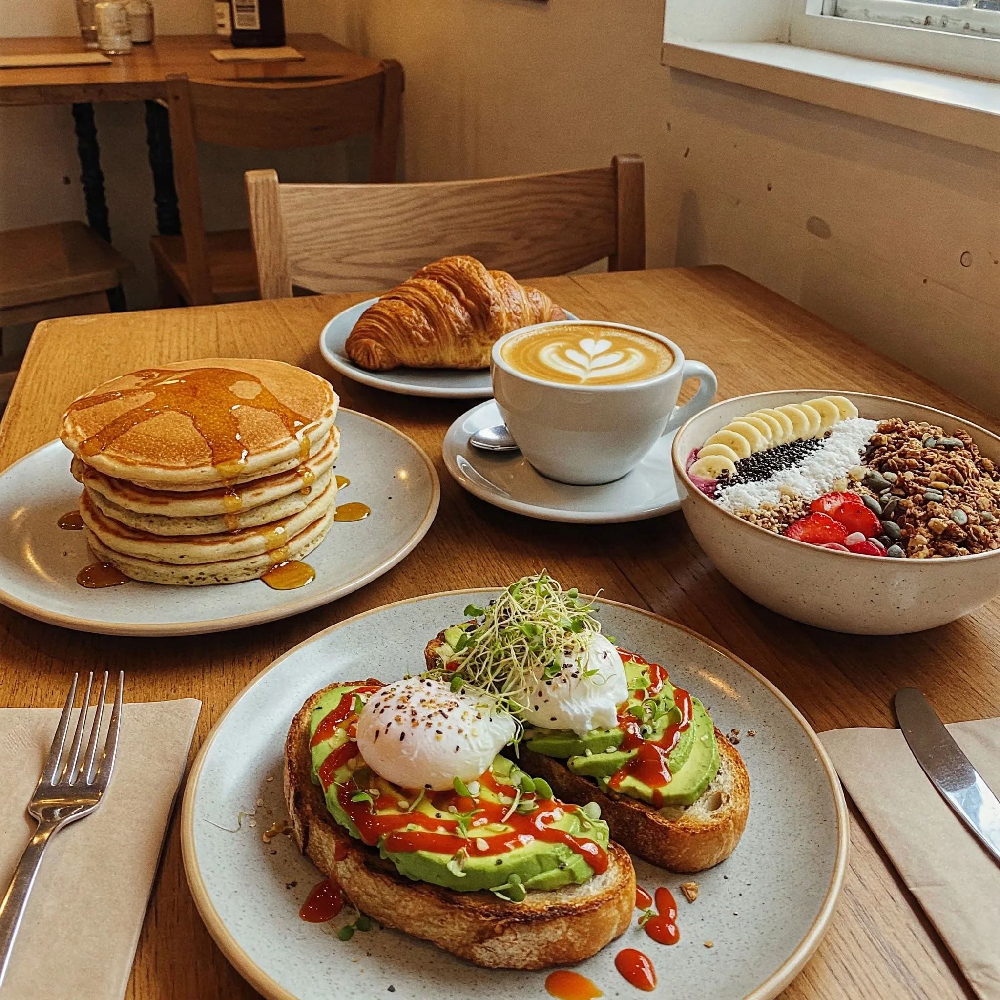 A variety of delicious breakfast items on a rustic wooden table, including pancakes, avocado toast, a smoothie bowl, coffee, and a croissant, set in a cozy café atmosphere.

