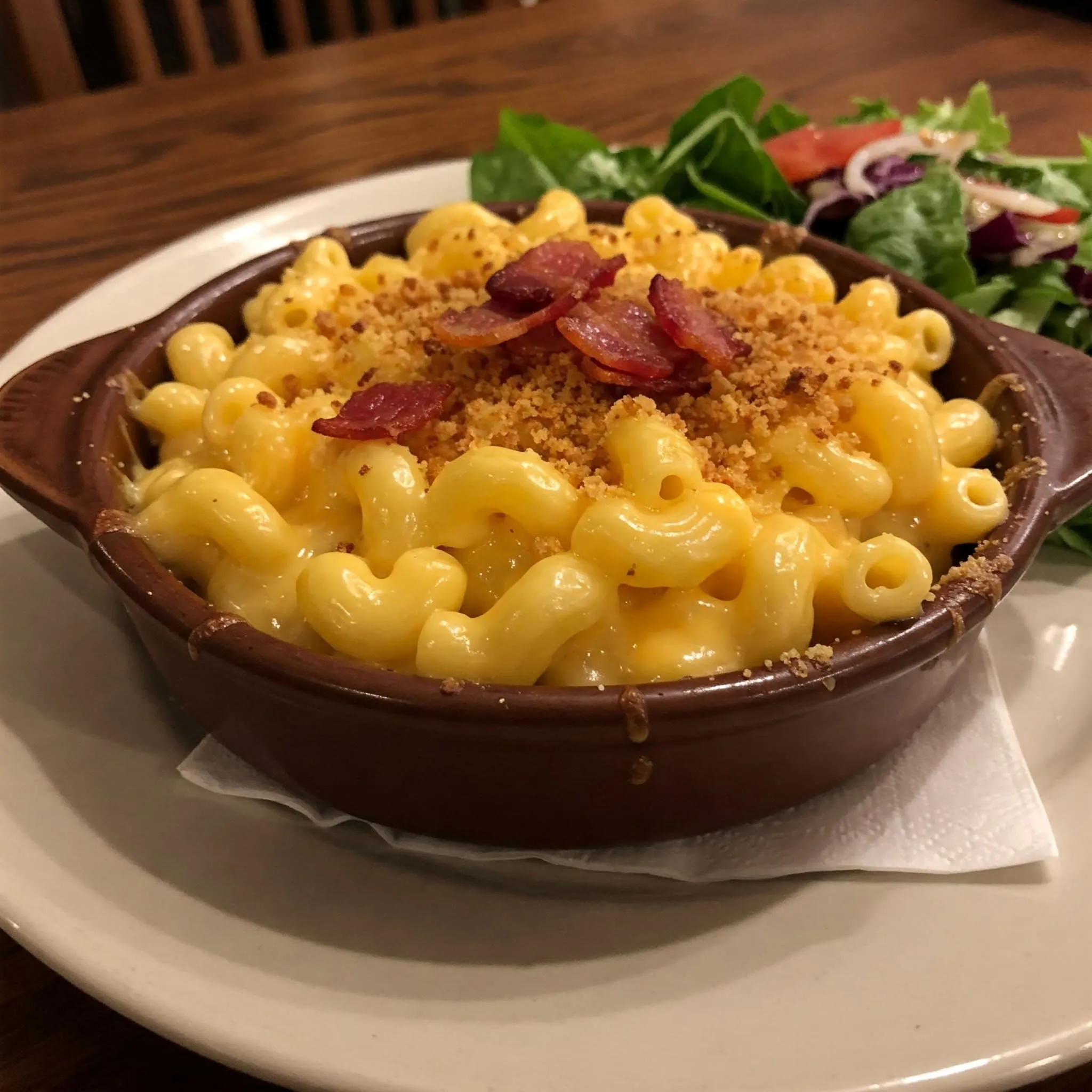 A bowl of creamy mac and cheese topped with crispy breadcrumbs and bacon pieces, served with a fresh side salad.