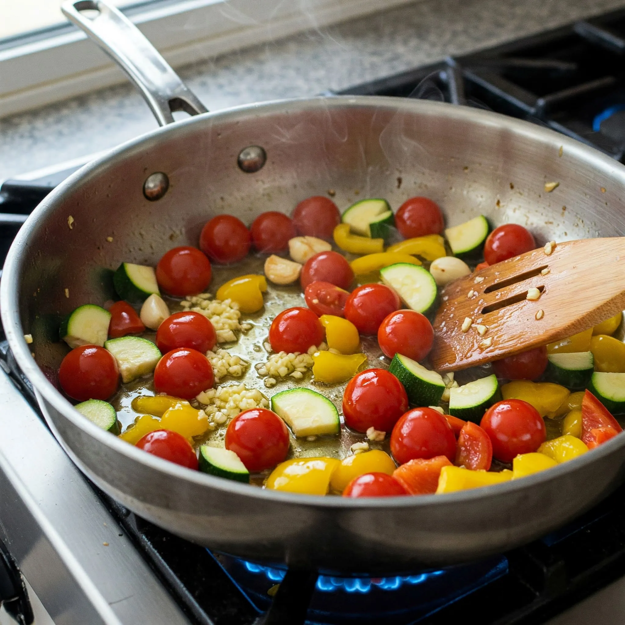 Sautéed vegetables including garlic, cherry tomatoes, zucchini, and bell pepper in olive oil, sizzling in a large pan on the stove.